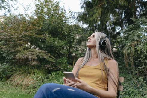 Frau mit Handy sitzt im Garten und hört Musik mit Kopfhörern - KNSF03540