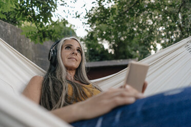 Portrait of woman with cell phone lying in hammock in the garden listening music with headphones - KNSF03541