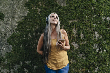 Frau mit Handy steht vor einer Wand und hört Musik mit Kopfhörern - KNSF03543