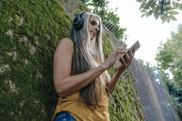 Frau mit Handy steht im Garten und hört Musik mit Kopfhörern - KNSF03544