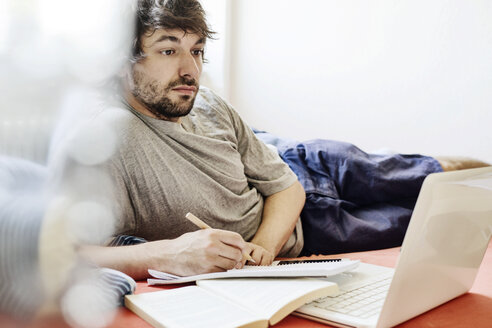 Portrait of student lying on bed with laptop and book learning - JATF00974