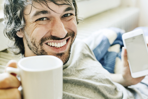 Porträt eines lachenden jungen Mannes auf dem Bett liegend mit Smartphone und Tasse Kaffee, lizenzfreies Stockfoto