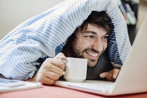 Portrait of laughing young man lying on bed with cup of coffee using laptop - JATF00976