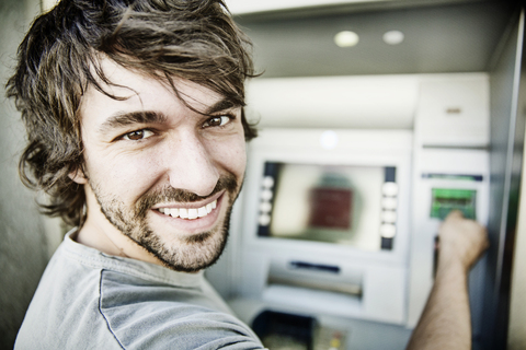 Porträt eines lachenden jungen Mannes an einem Geldautomaten, lizenzfreies Stockfoto