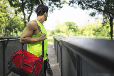 Runner standing on bridge in park, carrying bag and shoes - SBOF01135