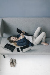 Businesswoman lying on the couch with several electronic devices using tablet - JOSF02098
