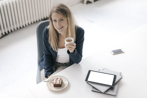 Porträt einer lächelnden Geschäftsfrau bei Kaffee und Kuchen am Schreibtisch im Büro, lizenzfreies Stockfoto