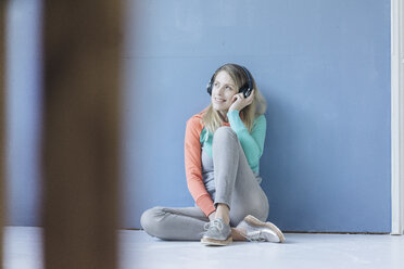 Portrait of smiling woman sitting on the floor in front of blue wall listening music with wireless headphones - JOSF02110
