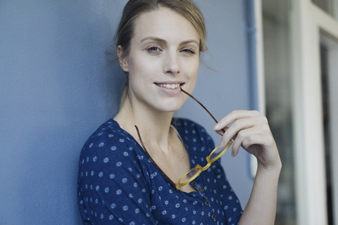 Porträt einer lächelnden Frau mit Brille, die sich an eine blaue Wand lehnt, lizenzfreies Stockfoto