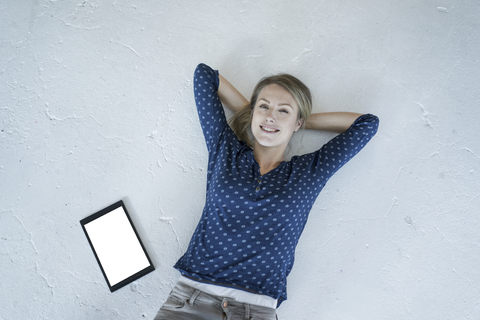 Porträt einer lächelnden jungen Frau, die mit einem Tablet auf dem Boden liegt, lizenzfreies Stockfoto