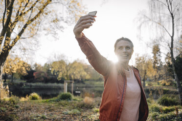 Porträt einer jungen Frau, die ein Selfie mit ihrem Handy in einem herbstlichen Park macht - JOSF02154