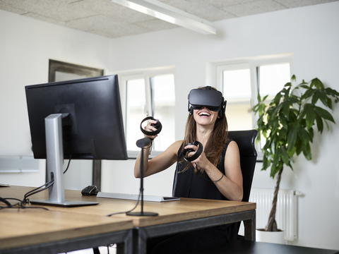 Glückliche Frau mit VR-Brille am Schreibtisch im Büro, lizenzfreies Stockfoto