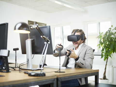 Glücklicher Mann mit VR-Brille am Schreibtisch im Büro, lizenzfreies Stockfoto