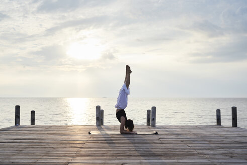 Young woman practicing yoga on a jetty by the sea at sunset - IGGF00390