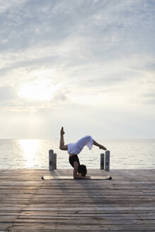 Junge Frau übt Yoga auf einem Steg am Meer bei Sonnenuntergang - IGGF00391