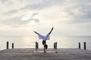 Junge Frau übt Yoga auf einem Steg am Meer bei Sonnenuntergang - IGGF00392