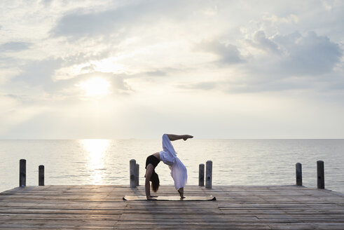 Junge Frau übt Yoga auf einem Steg am Meer bei Sonnenuntergang - IGGF00393