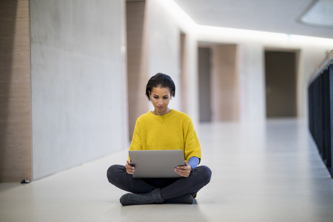 Porträt einer jungen Frau mit gelbem Pullover, die auf dem Boden sitzt und einen Laptop benutzt, lizenzfreies Stockfoto