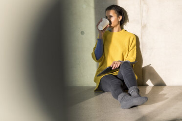 Junge Frau in gelbem Pullover sitzt auf dem Boden und trinkt Kaffee zum Mitnehmen - FMKF04712