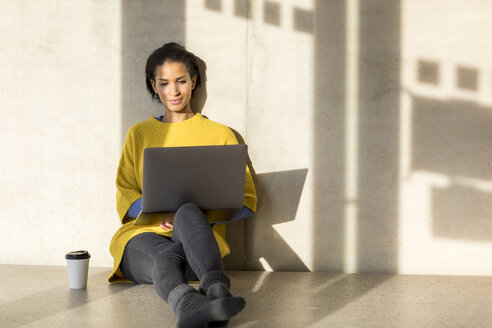 Porträt einer lächelnden jungen Frau, die auf dem Boden sitzt und einen Laptop benutzt - FMKF04713