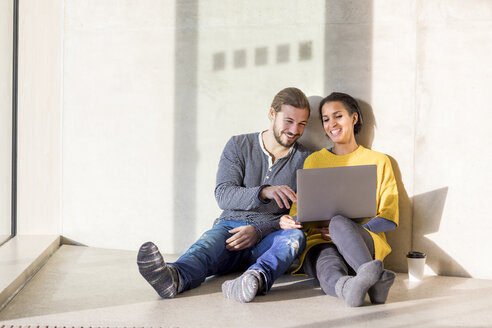 Porträt eines lachenden jungen Paares, das auf dem Boden sitzt und einen Laptop benutzt - FMKF04715
