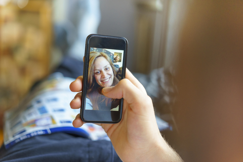 Porträt einer glücklichen jungen Frau auf dem Display eines Handys, die mit ihrem auf der Couch liegenden Freund skyped, lizenzfreies Stockfoto