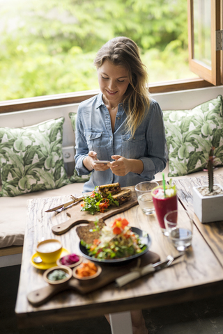 Frau fotografiert Essen mit Smartphone in einem gemütlichen Café vor einem Fenster, lizenzfreies Stockfoto