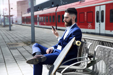Junger Mann mit Longboard sitzt am Bahnhof und benutzt sein Smartphone - MAEF12502