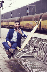 Junger Mann mit Longboard sitzt am Bahnhof und benutzt sein Smartphone - MAEF12503