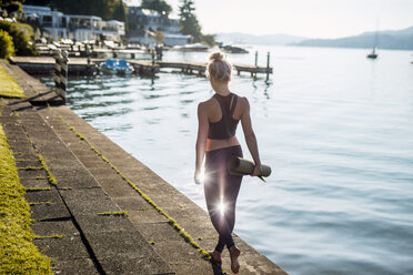 Rear view of woman in sportswear walking with yoga mat at lakeshore - DAWF00583