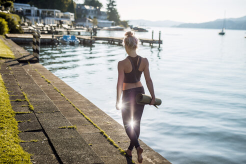 Rückansicht einer Frau in Sportkleidung, die mit einer Yogamatte am Seeufer spazieren geht - DAWF00583