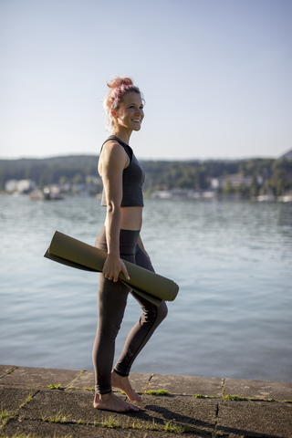 Woman in sportswear walking with yoga mat at lakeshore stock photo