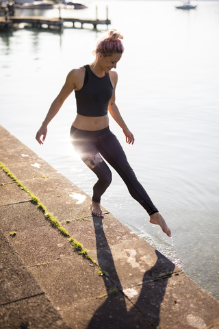 Frau in Sportkleidung am Seeufer hält Fuß ins Wasser, lizenzfreies Stockfoto