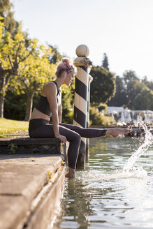 Frau in Sportkleidung am Seeufer sitzend mit den Füßen im Wasser - DAWF00588