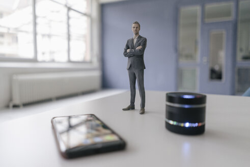 Miniature businessman figurine standing next to smart home loudspeaker and smartphone - FLAF00126