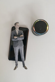 Miniature businessman figurine lying on smartphone next to smart home loudspeaker - FLAF00128