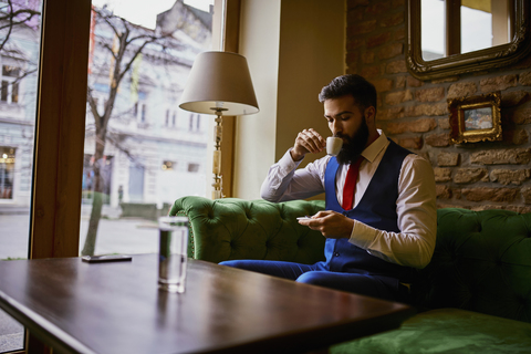 Fashionable young man sitting on couch in a cafe drinking coffee and using cell phone stock photo