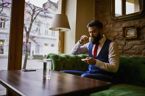 Modischer junger Mann sitzt auf der Couch in einem Café, trinkt Kaffee und benutzt sein Handy - ZEDF01100
