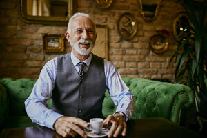 Porträt eines eleganten älteren Mannes, der lächelnd auf einer Couch in einem Café sitzt - ZEDF01105