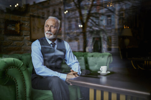 Elegant senior man sitting on couch in a cafe looking out of window - ZEDF01111