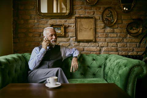 Eleganter älterer Mann sitzt auf der Couch in einem Café und spricht mit seinem Handy, lizenzfreies Stockfoto