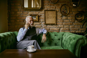 Eleganter älterer Mann sitzt auf der Couch in einem Café und spricht mit seinem Handy - ZEDF01113
