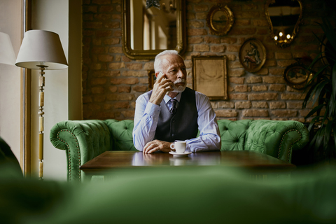 Elegant senior man sitting on couch in a cafe on cell phone stock photo