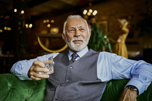 Portrait of elegant senior man sitting on couch in a bar holding tumbler - ZEDF01119