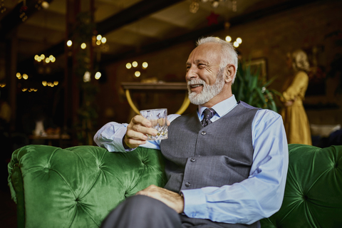 Porträt eines eleganten älteren Mannes, der auf einer Couch in einer Bar sitzt und einen Becher hält, lizenzfreies Stockfoto