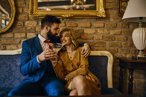 Glückliches elegantes Paar mit Getränken auf der Couch sitzend und sich umarmend - ZEDF01164