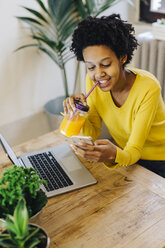 Young woman sitting at table, drinking juice and checking smartphone - GIOF03800