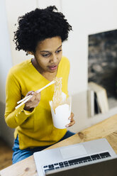 Young woman sitting at table, surfing the web on laptop while eating noodles - GIOF03818