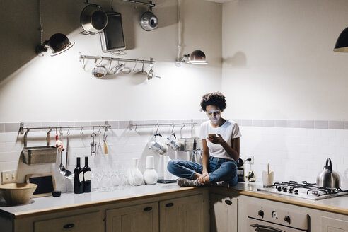 Young woman sitting on kitchen worktop, checking messages on smartphone - GIOF03842