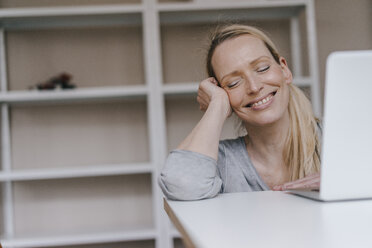 Smiling woman with closed eyes leaning on table with laptop - KNSF03573